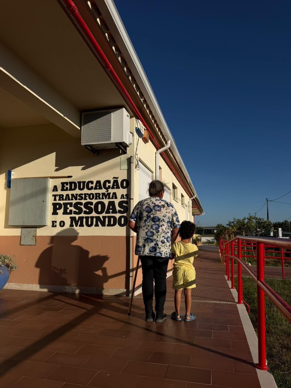 Uma senhora de bengala e uma criança de mão dada à entrada do Centro Social, junto à frase A educação transforma as pessoas e o mundo